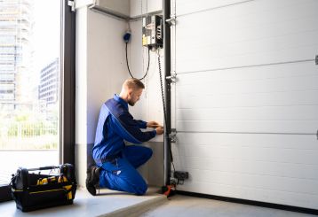 Technician in blue uniform kneeling beside a newly installed white garage door, adjusting the opener system and hardware in a Cartersville, GA home garage.