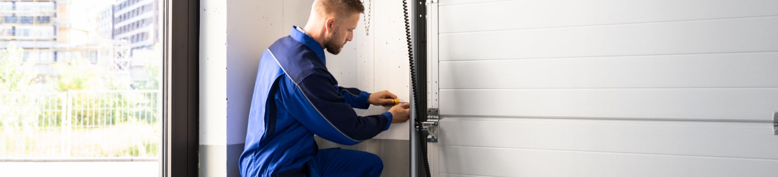 Technician in blue uniform kneeling beside a newly installed white garage door, adjusting the opener system and hardware in a Cartersville, GA home garage.